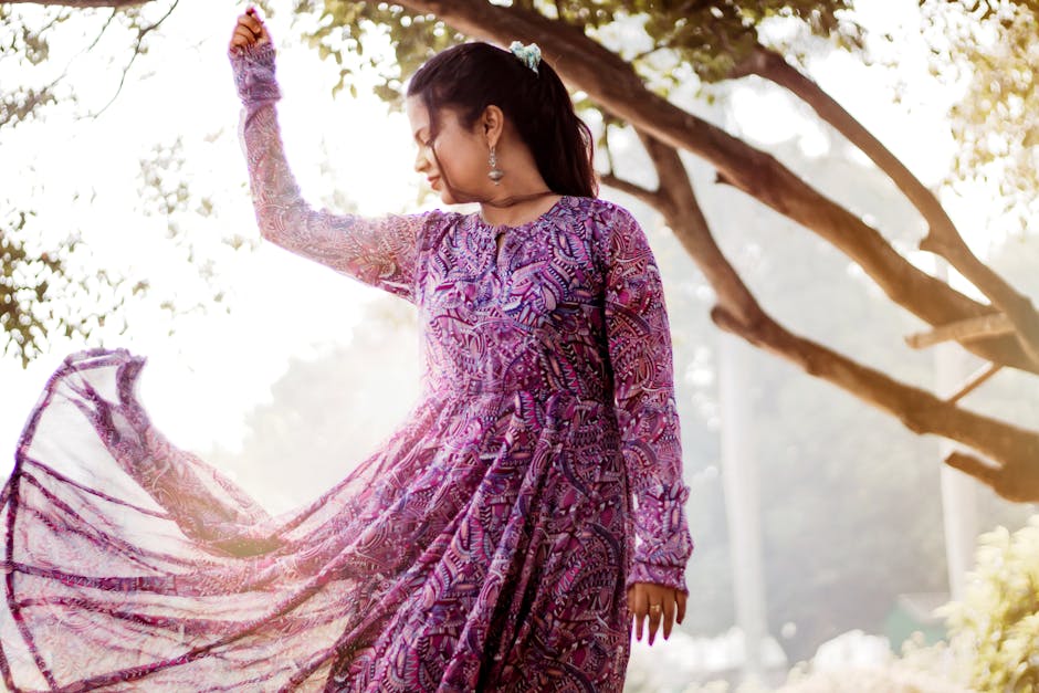 A woman poses gracefully in a vibrant purple dress, basking in sunlight amidst the trees.