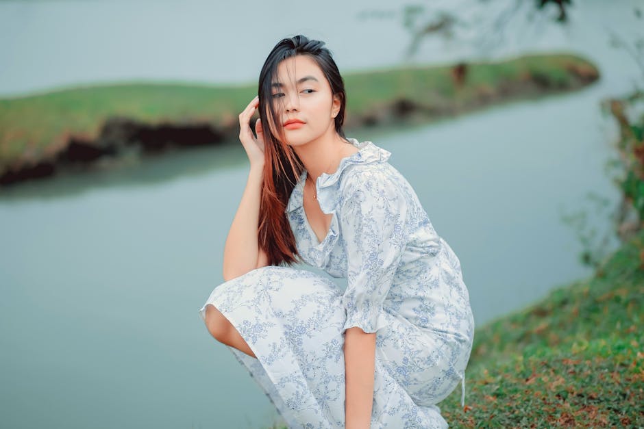 Graceful woman in a floral dress crouching by a tranquil riverbank in summer.