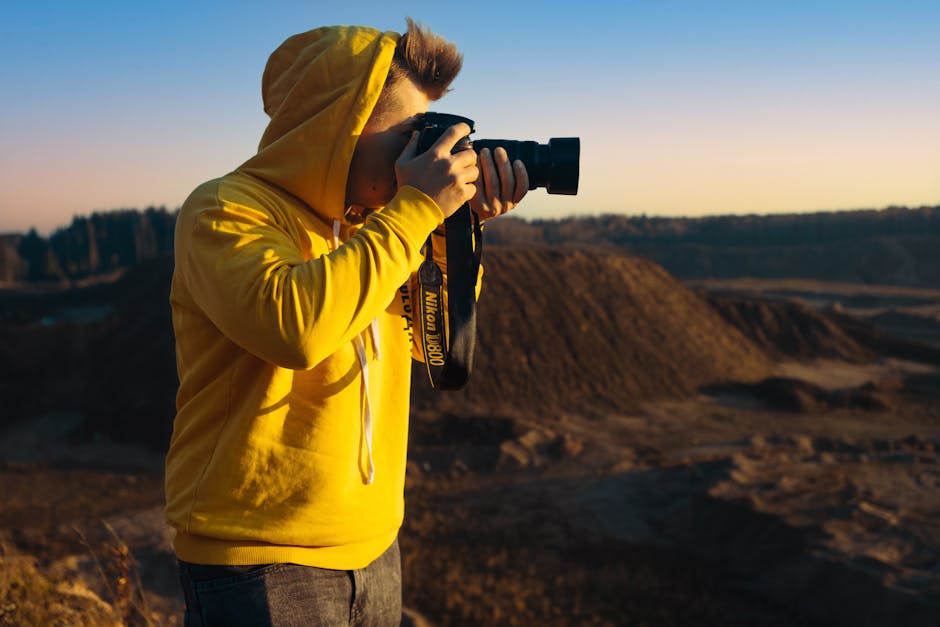 A young adult in a yellow hoodie photographs a scenic valley during sunset.