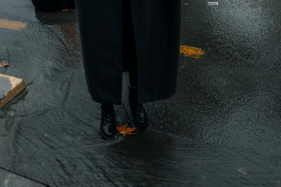 A person in a long coat stands on a wet street with fallen autumn leaves.