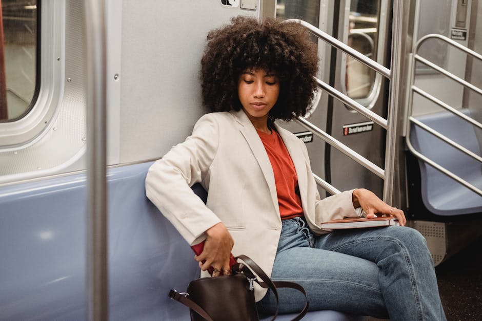 A woman in a white blazer sitting on a subway train, holding a notebook and red shirt visible.