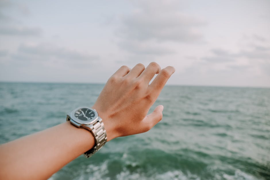 A close-up shot of a hand wearing a wristwatch, reaching towards the ocean under a cloudy sky.