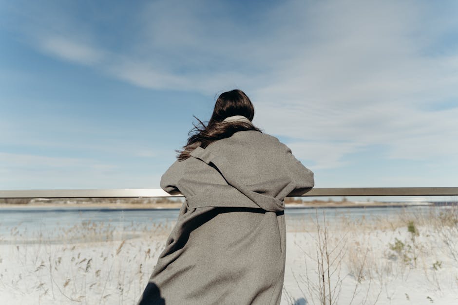 A woman in a gray coat stands on a terrace, gazing at a snowy landscape under a blue sky.
