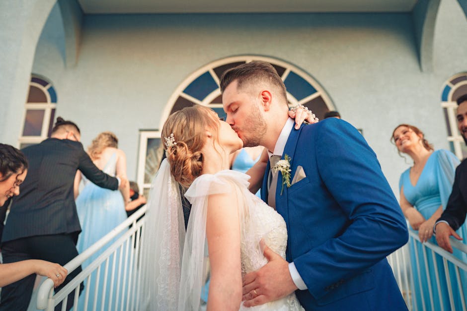 A couple shares a romantic kiss at their wedding, surrounded by guests in formal attire.
